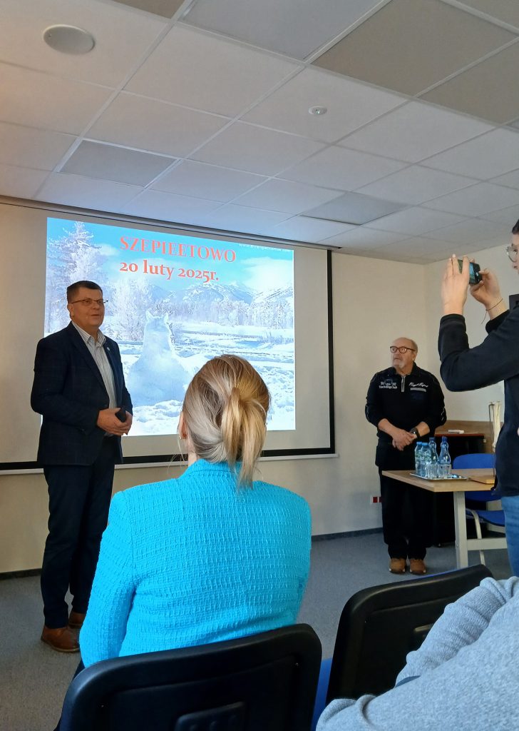 W pomieszczeniu znajduje się kilku uczestników konferencji – na pierwszym planie widać kobietę w niebieskiej marynarce siedzącą na krześle, a obok niej innych słuchaczy.

Na tle ekranu z prezentacją stoi mężczyzna w garniturze, który prawdopodobnie wygłasza przemówienie. Obok niego znajduje się starszy mężczyzna ubrany na czarno, który wygląda na jednego z organizatorów lub ekspertów. Po prawej stronie ktoś robi zdjęcie aparatem.