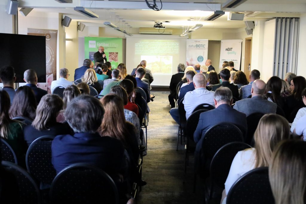 Na zdjęciu widać konferencję lub prezentację odbywającą się w sali konferencyjnej. Uczestnicy siedzą na czarnych, tapicerowanych krzesłach ustawionych w rzędach, zwróceni w stronę prelegenta, który stoi przy ekranie i mówi do mikrofonu. Na ekranie wyświetlana jest prezentacja z tekstem i obrazem, a za mówcą znajdują się banery reklamowe lub informacyjne z napisami, takimi jak „Fortibac Plus” i „naturBIOTIC”. Sala jest dobrze oświetlona, a publiczność składa się z ludzi w różnym wieku, ubranych w stroje biznesowe lub casualowe.