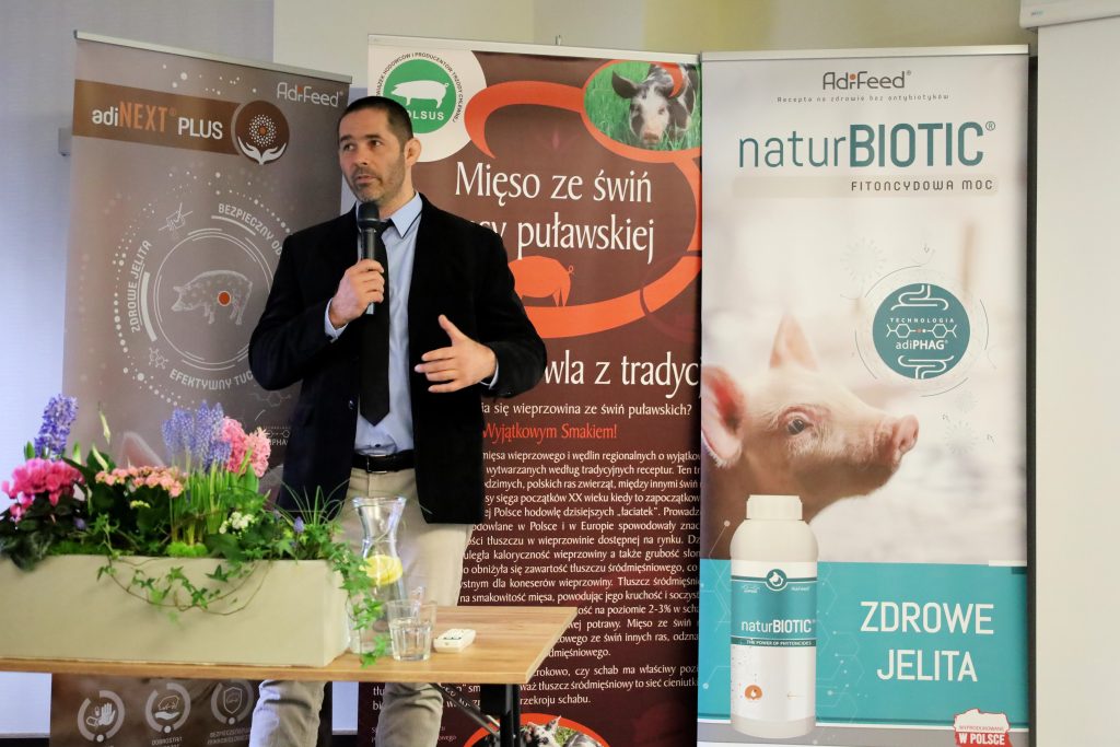 Na zdjęciu widzimy mężczyznę w garniturze, który trzyma mikrofon i wygłasza prezentację. Stoi on przed trzema roll-upami reklamowymi, które promują produkty związane z hodowlą świń i zdrowiem zwierząt.