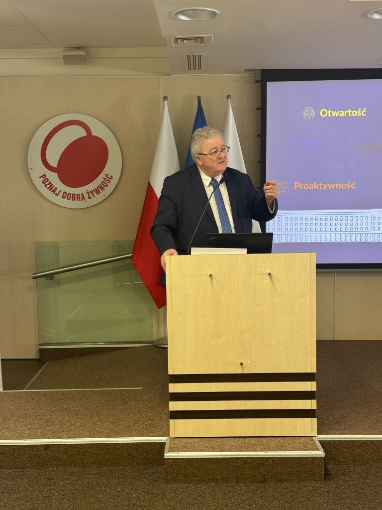 Na zdjęciu mężczyzna w garniturze przemawia zza mównicy w sali konferencyjnej. W tle widać ekran z prezentacją, na którym widnieją hasła „Otwartość” i „Proaktywność”, a także flagi Polski i Unii Europejskiej. Po lewej stronie widnieje logo programu „Poznaj Dobrą Żywność”.