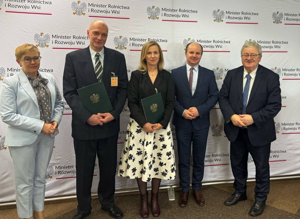 Grupa sześciu osób pozuje do zdjęcia na tle ścianki Ministerstwa Rolnictwa i Rozwoju Wsi. Dwie osoby trzymają teczki z godłem, co sugeruje wręczenie nagród lub wyróżnień.