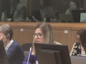Kobieta w okularach mówi do mikrofonu podczas konferencji, skupiona na wypowiedzi. W tle inni uczestnicy pracują przy komputerach i słuchają obrad.