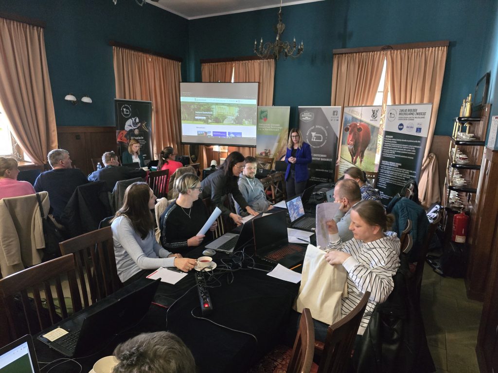 Konferencja w sali z ciemnozielonymi ścianami. Uczestnicy przy stole z laptopami i dokumentami. Kobieta prowadzi prezentację, w tle banery rolnicze i hodowlane.