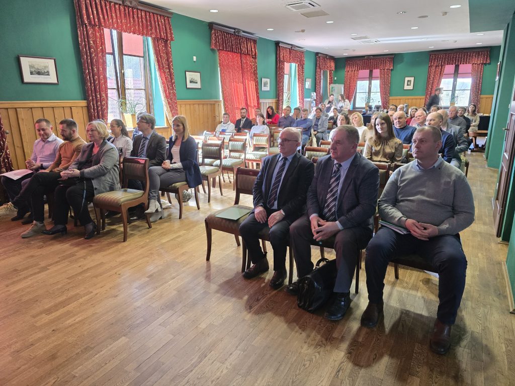 Uczestnicy konferencji siedzą w sali z zielonymi ścianami i czerwonymi zasłonami. W pierwszych rzędach widoczne są osoby w eleganckich strojach, a w tle kolejni uczestnicy skupieni na przebiegu wydarzenia.