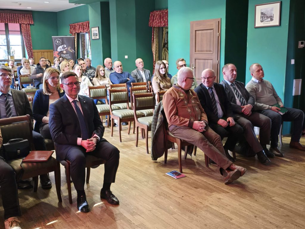 Uczestnicy konferencji siedzą w rzędach w sali o zielonych ścianach. W pierwszym rzędzie kilku mężczyzn w garniturach i eleganckich ubraniach prowadzi rozmowę, a pozostali słuchają przebiegu wydarzenia.