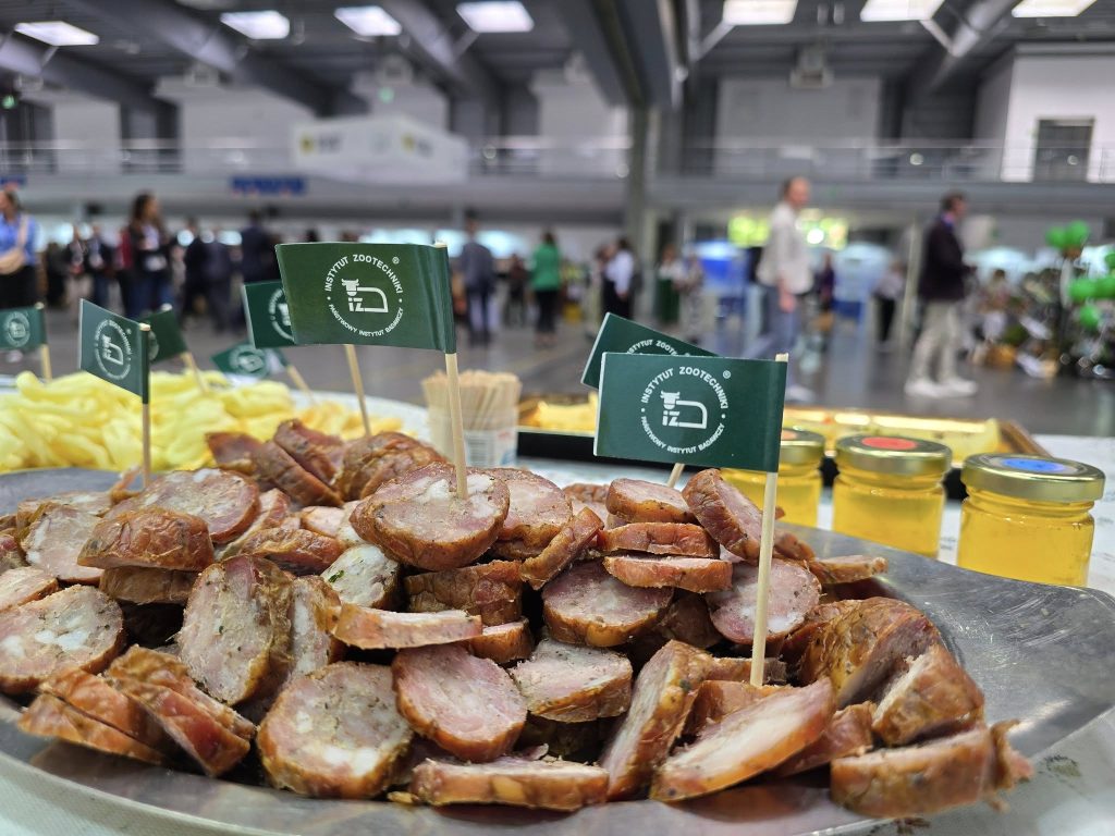 Stół degustacyjny z próbkami żywności oznaczonymi flagami Instytutu Zootechniki; w tle uczestnicy wydarzenia i stoiska wystawowe w przestrzeni targowej.