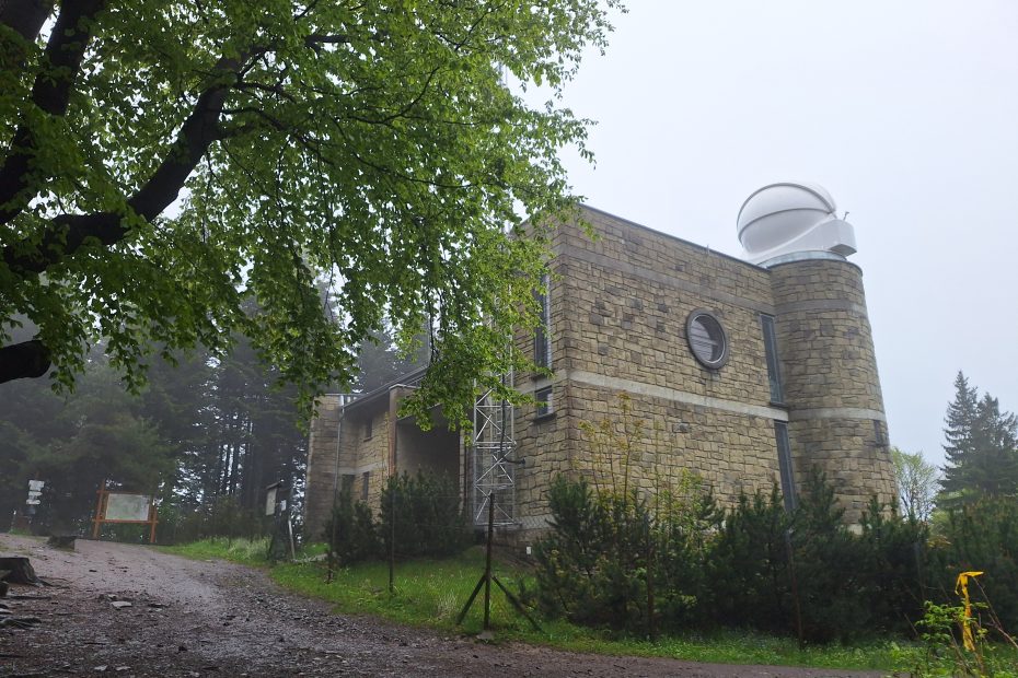 Kamienny budynek Obserwatorium Astronomicznego na Lubomirze otoczony zielenią i lekką mgłą; na dachu widoczna biała kopuła teleskopu, a w tle drzewa i tablica informacyjna przy leśnym szlaku.