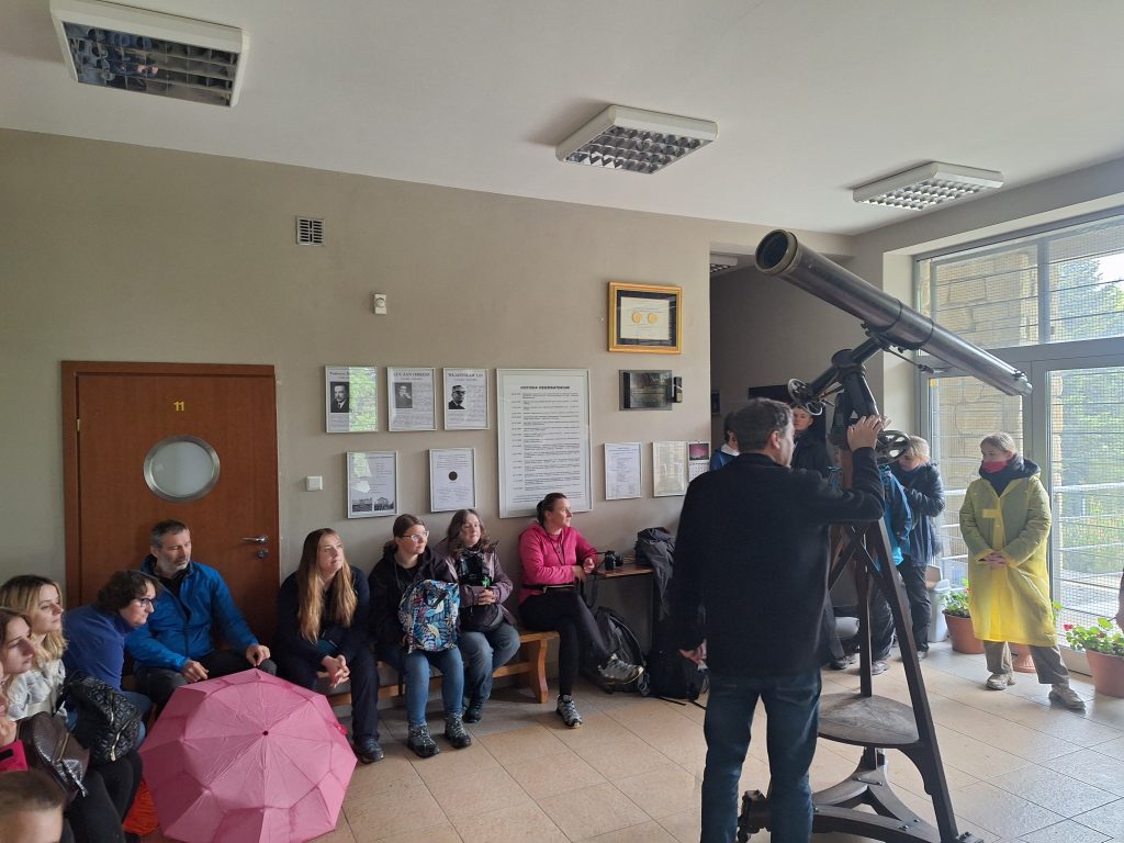 Grupa uczestników Rajdu Cielaka słucha prelekcji wewnątrz Obserwatorium Astronomicznego na Lubomirze; na pierwszym planie prowadzący opiera rękę na zabytkowym teleskopie, w tle widoczne tablice informacyjne i okno z widokiem na zewnątrz.