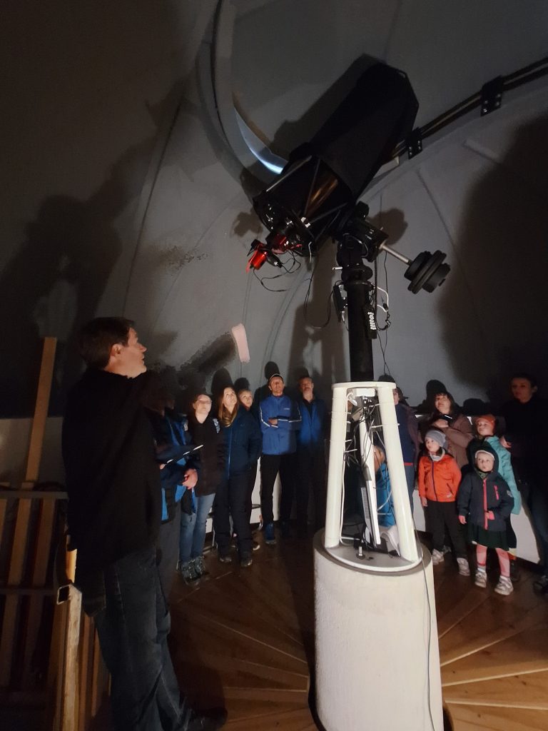 Wnętrze kopuły Obserwatorium Astronomicznego na Lubomirze. Grupa uczestników, w tym dzieci, zgromadziła się wokół nowoczesnego teleskopu, słuchając przewodnika objaśniającego zasady jego działania. Atmosferę skupienia podkreśla przyciemnione światło i surowe, techniczne wnętrze.