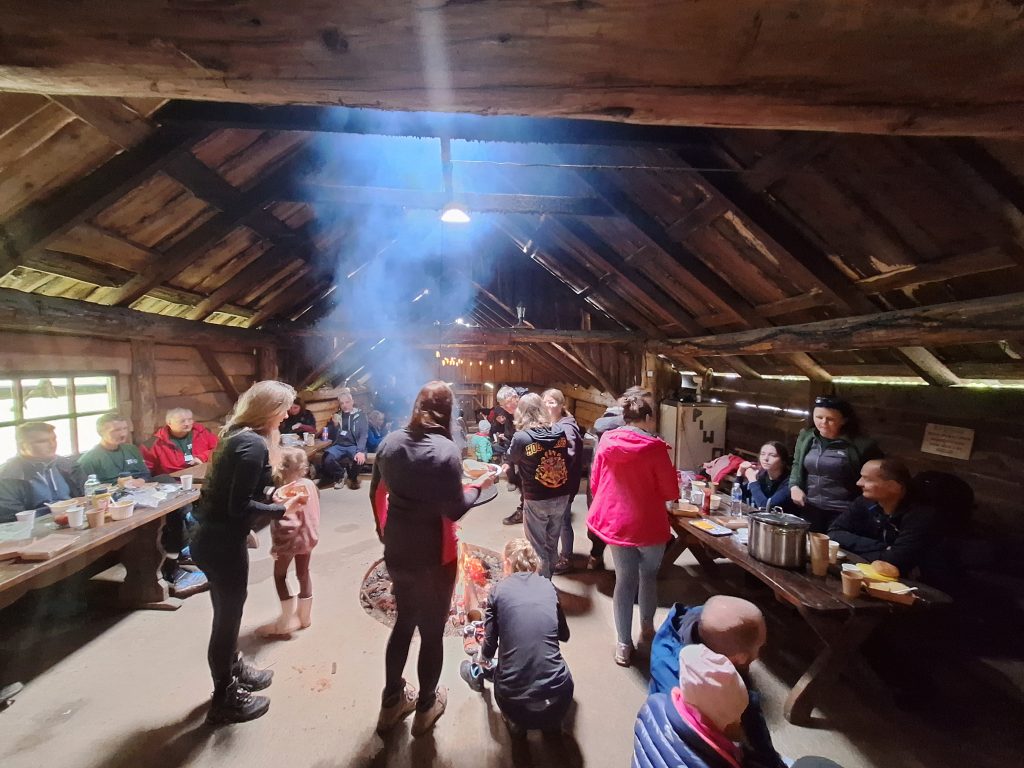 Uczestnicy rajdu siedzą i stoją przy ognisku w drewnianym szałasie, jedząc posiłek i rozmawiając.
