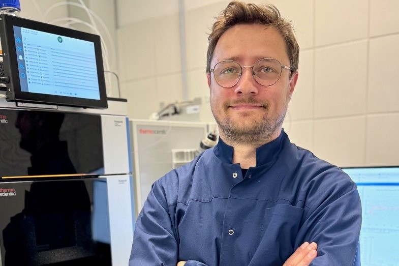 Mężczyzna w okularach i niebieskim kitlu laboratoryjnym stoi z założonymi rękami w nowoczesnym laboratorium.