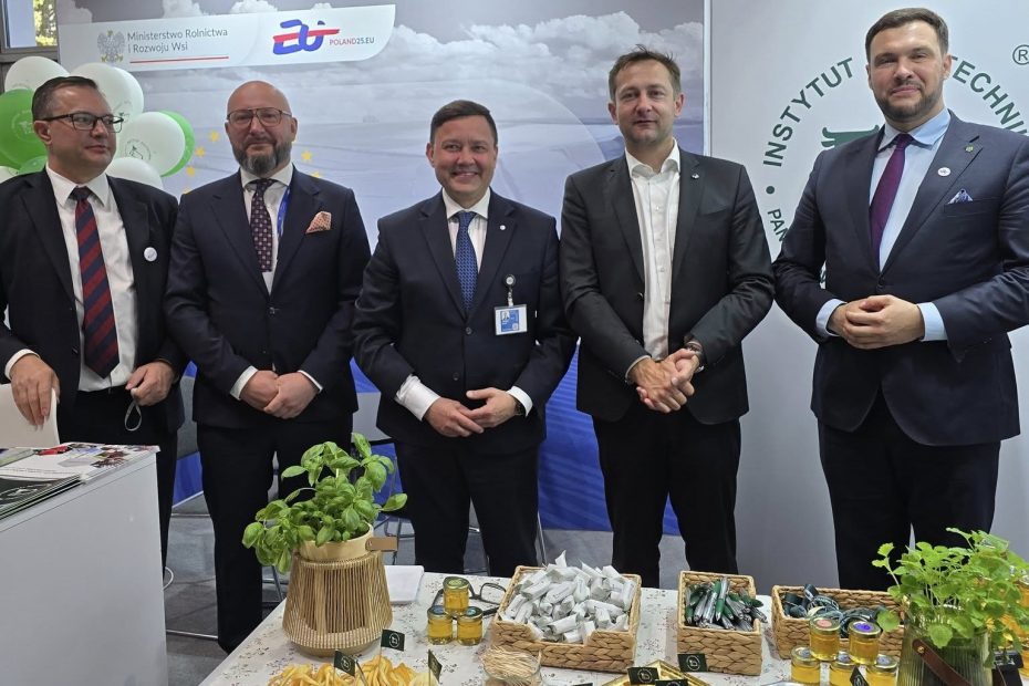 Delegacja mężczyzn w garniturach stojąca przy stoisku Instytutu Zootechniki podczas wydarzenia promującego polskie rolnictwo; w tle logo Ministerstwa Rolnictwa i Rozwoju Wsi oraz symbol 20-lecia Polski w UE.