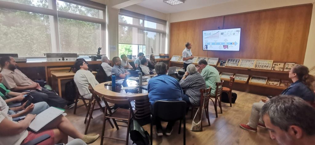 Spotkanie robocze w sali entomologicznej – uczestnicy siedzą przy stołach i słuchają prezentacji mężczyzny przy ekranie, na którym widnieje slajd z wykresami i schematem. W tle gabloty z owadami.