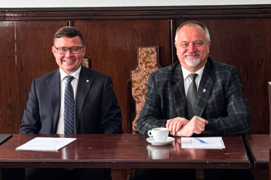 Dwaj uśmiechnięci mężczyźni w garniturach siedzą przy stole podczas posiedzenia Rady Uczelni Uniwersytetu Rolniczego w Krakowie. Przed nimi leżą dokumenty i filiżanka z kawą