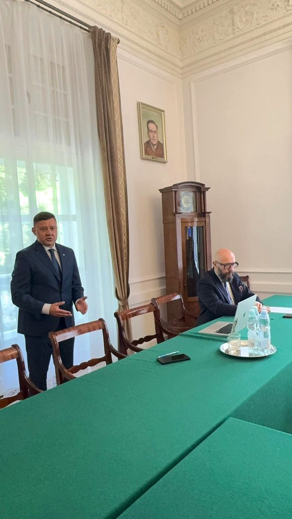 Dwóch mężczyzn w garniturach podczas spotkania w eleganckiej sali z zielonym stołem konferencyjnym; jeden stoi i mówi, drugi siedzi przy laptopie. W tle zabytkowy zegar i portret na ścianie.