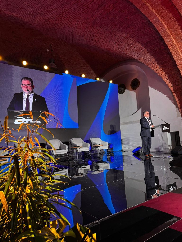 Mężczyzna w garniturze przemawia na scenie przy mównicy z logo „Polska 2025”; za nim ekran z jego wizerunkiem i kolorowe oświetlenie. Scena znajduje się w sali z ceglanym sklepieniem i kilkoma krzesłami ustawionymi z boku.