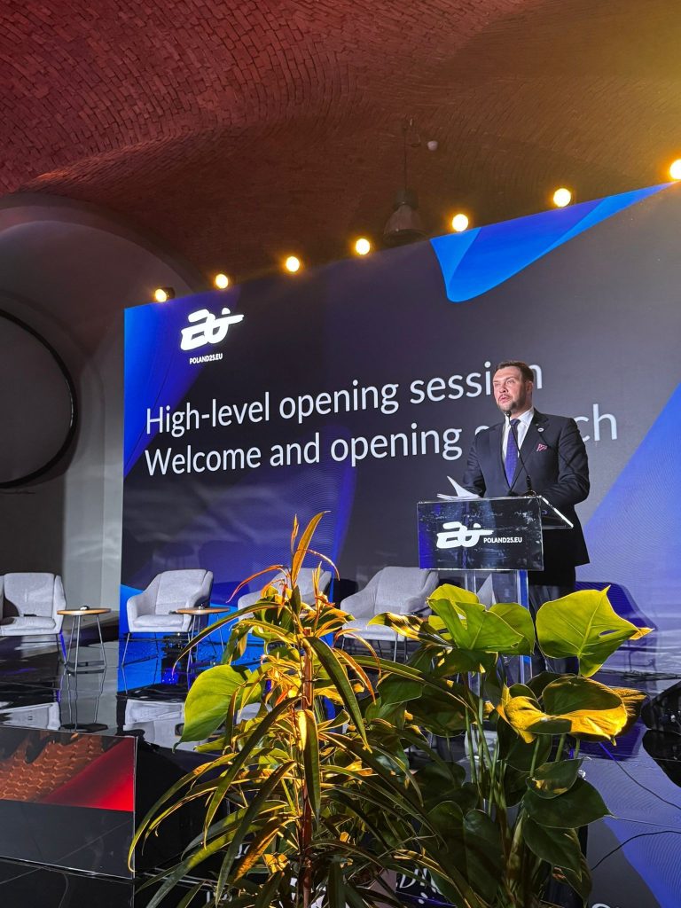 Mężczyzna w garniturze przemawia na scenie przy mównicy z logo „Poland 2025”, na tle dużego ekranu z napisem „High-level opening session – Welcome and opening speech”. W tle ustawiono kilka krzeseł, a na pierwszym planie widoczne są rośliny ozdobne.