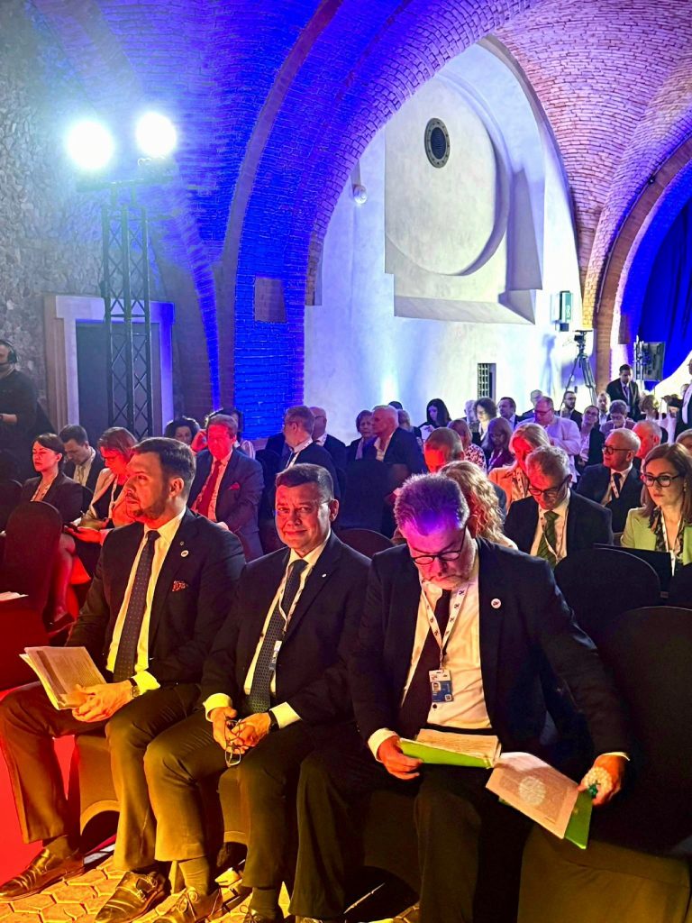 Uczestnicy wydarzenia siedzą w rzędach na sali z ceglanym sklepieniem, oświetlonej na niebiesko i czerwono. W pierwszym rzędzie widać kilku mężczyzn w garniturach, z identyfikatorami i dokumentami w rękach. W tle pozostała część publiczności i ekipa techniczna.