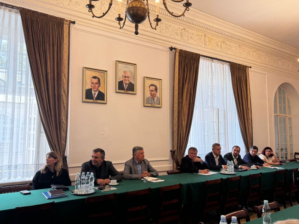 Grupa osób siedzi przy zielonym stole konferencyjnym w eleganckiej sali z wysokimi oknami i zasłonami; na ścianie wiszą portrety trzech mężczyzn. Uczestnicy spotkania notują i rozmawiają.