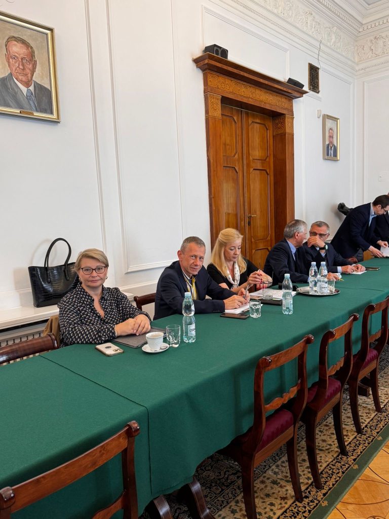 Uczestnicy spotkania siedzą przy zielonym stole konferencyjnym w eleganckiej sali z portretami na ścianach i drewnianymi drzwiami. Na stole stoją butelki wody, filiżanki i notatniki.