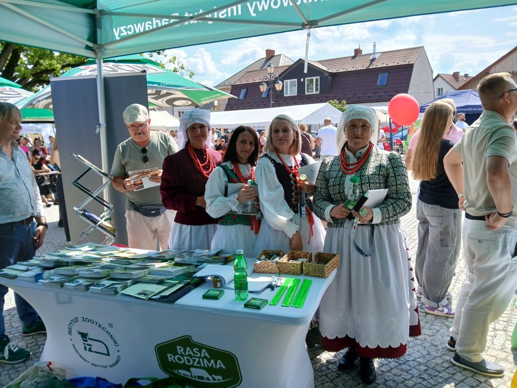 Kobiety w tradycyjnych strojach ludowych stoją przy stoliku promocyjnym Instytutu Zootechniki – PIB pod zielonym namiotem. Na stole ulotki, gadżety i woda. W tle widoczni uczestnicy wydarzenia i inne stoiska.