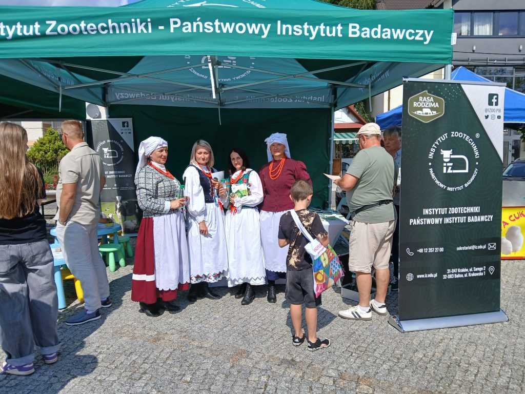 Stoisko Instytutu Zootechniki z zielonym namiotem i banerami informacyjnymi. Przed stoiskiem kilka kobiet w tradycyjnych strojach ludowych oraz odwiedzający, w tym dziecko z kolorową torbą.