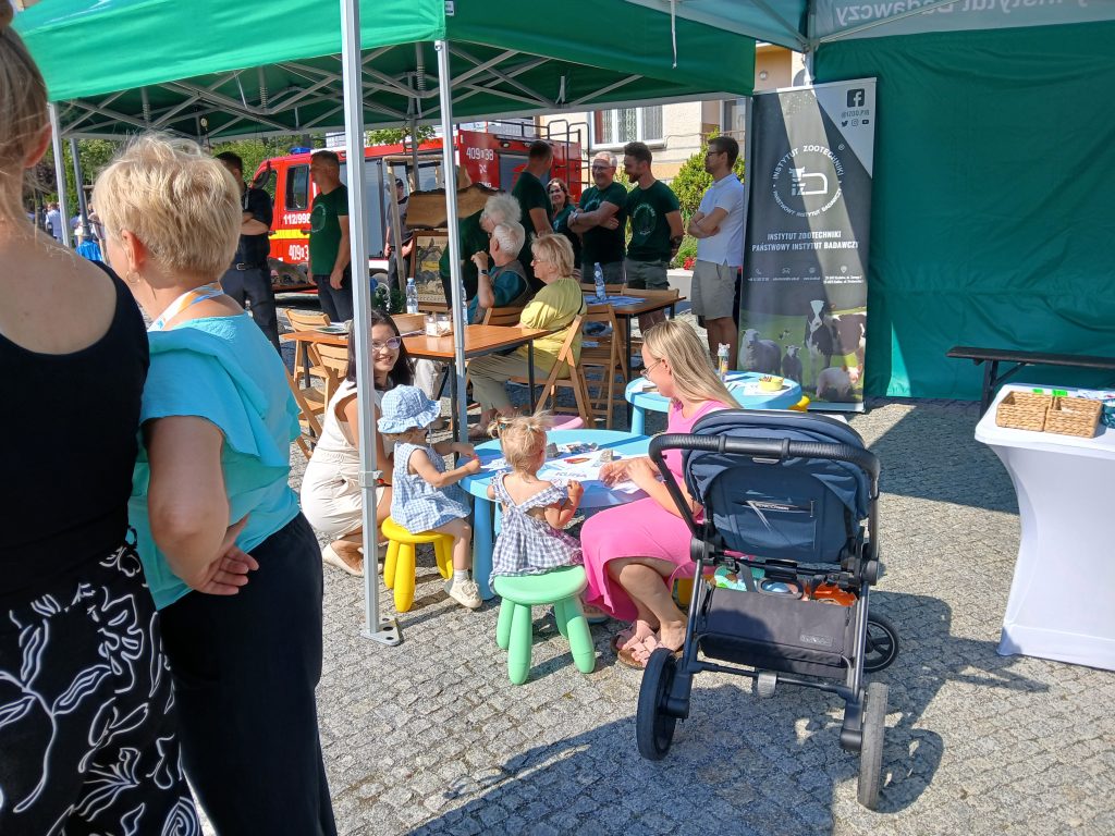 Dzieci uczestniczą w zabawie plastycznej przy kolorowych stolikach pod namiotem Instytutu Zootechniki – PIB. Obok siedzi opiekunka, w tle widoczni dorośli, baner instytutu oraz wózek dziecięcy.