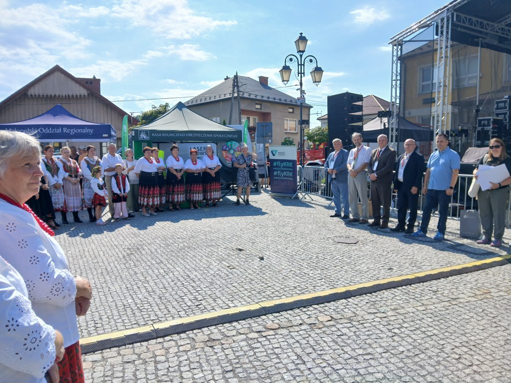 Uczestnicy wydarzenia ustawieni w dwóch rzędach na placu: po lewej stronie kobiety i dzieci w strojach ludowych, po prawej przedstawiciele władz i organizatorzy. W tle namioty promocyjne KRUS i Małopolskiego Oddziału Regionalnego