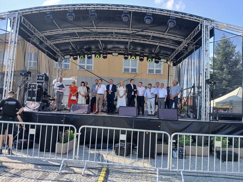 Grupa osób, w tym przedstawiciele władz i organizatorzy, stoi na scenie plenerowej podczas oficjalnego otwarcia wydarzenia. W tle budynek z banerem.
