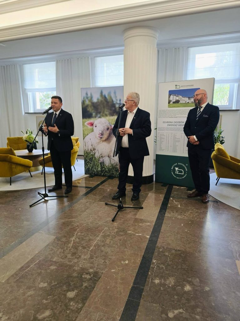 Dwaj mężczyźni przemawiający podczas otwarcia wystawy rodzimych ras zwierząt gospodarskich, w tle plakaty wystawowe.