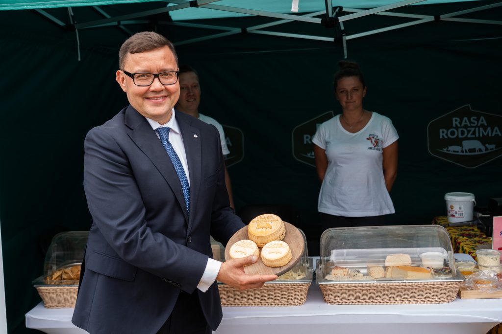 Mężczyzna w garniturze i krawacie prezentuje na drewnianej desce tradycyjne sery, stojąc przy stoisku z wyrobami mleczarskimi oznaczonym napisem „Rasa Rodzima”. W tle widoczne są osoby obsługujące stoisko.