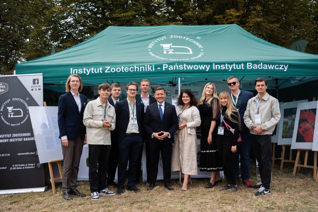 Grupa osób pozuje wspólnie do zdjęcia przed namiotem Instytutu Zootechniki – Państwowego Instytutu Badawczego. W tle widoczne są banery informacyjne oraz plansze ze zdjęciami zwierząt.