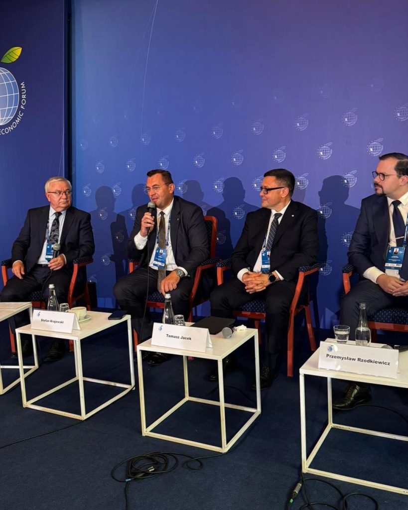 Czterech mężczyzn siedzi na scenie w fotelach, a przed nimi stoją stoły z wodą i mikrofonami. Jedna z osób (druga od lewej) mówi do mikrofonu.