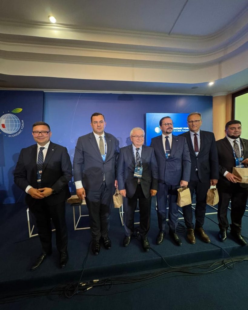 sześciu mężczyzn w garniturach stojących obok siebie na scenie podczas 34. Forum Ekonomicznego w Karpaczu (2–4 września 2025). Mają identyfikatory konferencyjne oraz trzymają papierowe torby upominkowe. W tle widoczne są ścianki z logo Forum Ekonomicznego i ekran informacyjny.