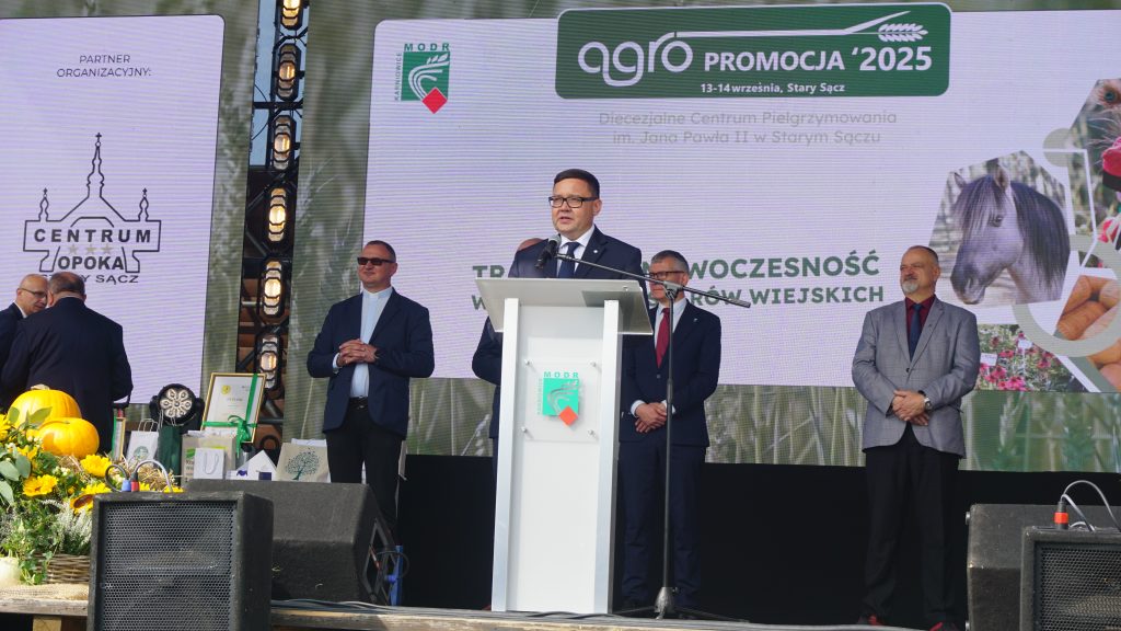 Mężczyzna w garniturze przemawia z mównicy na scenie podczas wydarzenia „Agro Promocja 2025” w Starym Sączu. Za nim stoją inni uczestnicy uroczystości, a w tle widoczny jest ekran z logotypami i hasłem o tradycji i nowoczesności na obszarach wiejskich.
