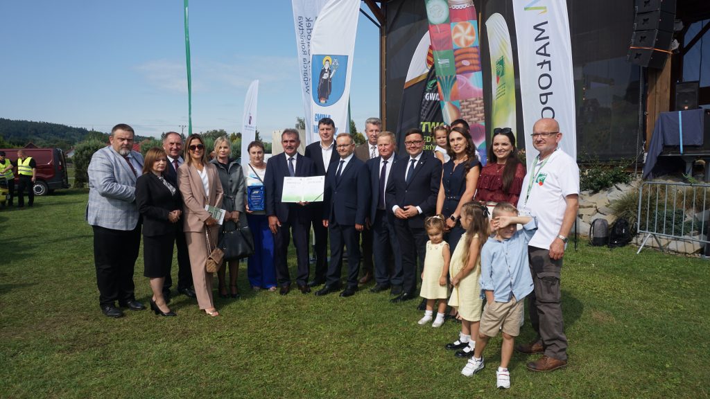 Grupa dorosłych i kilkoro dzieci pozuje do wspólnego zdjęcia na trawie podczas wydarzenia plenerowego „Agro Promocja 2025”. W tle widoczne są flagi z logotypami i fragment sceny.
