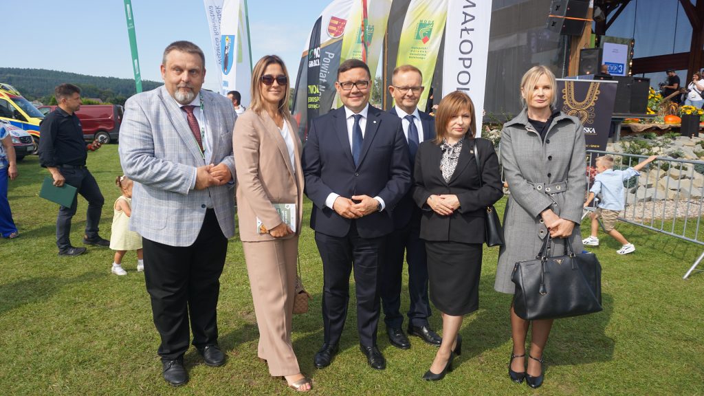 Grupa sześciu osób pozuje do zdjęcia na trawie podczas wydarzenia plenerowego „Agro Promocja 2025”. W tle widoczne są flagi z logotypami oraz fragment sceny.