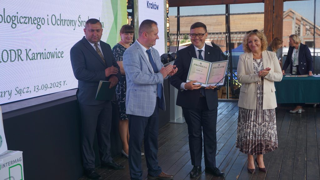 Mężczyzna w garniturze trzyma dyplom i promesę podczas uroczystości wręczenia nagród na scenie. Obok stoją inne osoby, niektórzy biją brawo, a w tle widoczny jest ekran z napisem o ODR Karniowice.