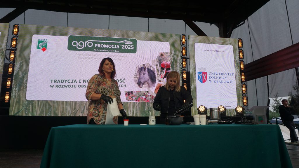 Dwie kobiety prowadzą pokaz na scenie wydarzenia „Agro Promocja 2025”, stojąc przy stole z naczyniami.