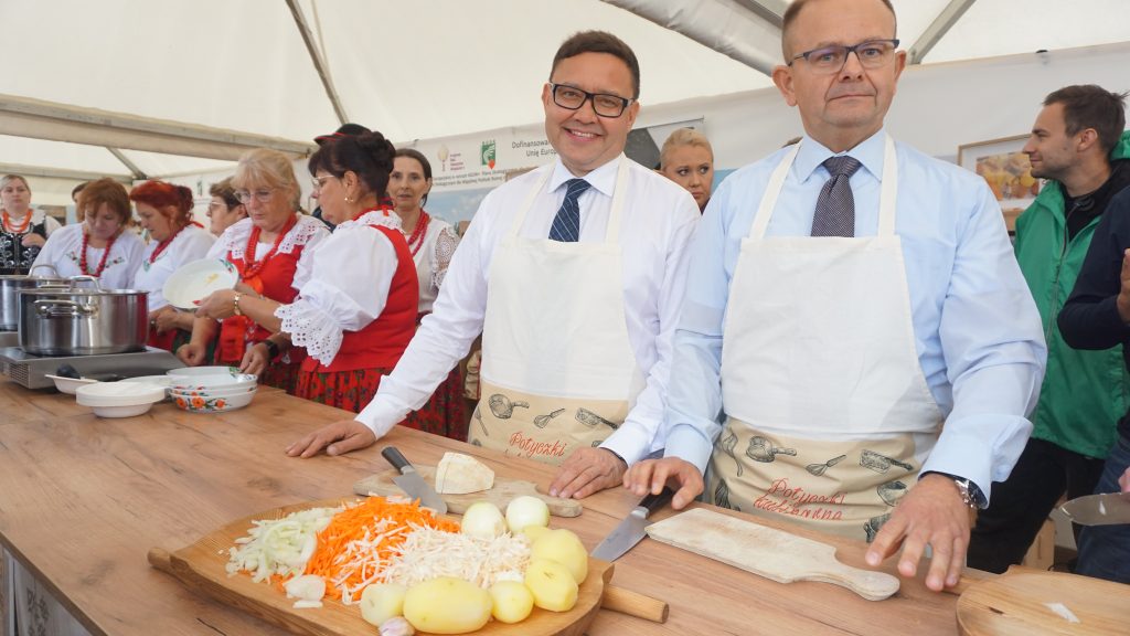 Dwaj mężczyźni w białych koszulach, krawatach i fartuchach kucharskich stoją przy stole z warzywami przygotowanymi do gotowania. W tle kobiety w strojach ludowych zajmują się kuchnią podczas pokazu kulinarnego.