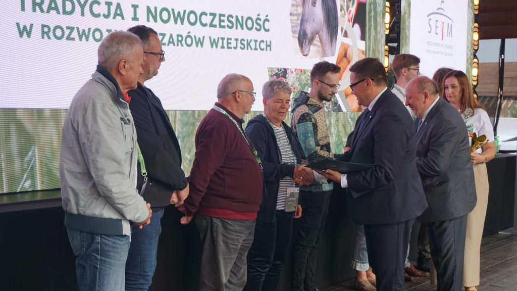 Uroczystość wręczenia nagród podczas wydarzenia „Agro Promocja 2025”. Mężczyzna w garniturze podaje dyplom i ściska dłoń jednej z osób stojących w kolejce po odbiór wyróżnień. W tle widoczny jest ekran z hasłem o tradycji i nowoczesności na obszarach wiejskich.