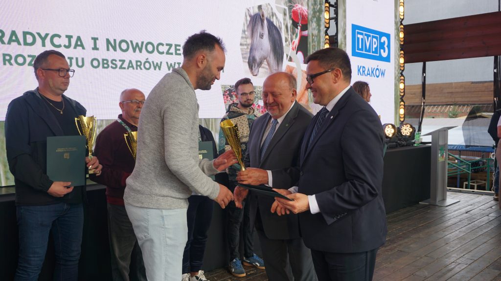 Wręczenie nagrody podczas wydarzenia „Agro Promocja 2025”. Mężczyzna w szarym swetrze odbiera złoty puchar od organizatorów w garniturach, a w tle stoją inni laureaci z trofeami i dyplomami.
