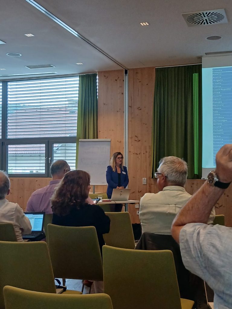 Salę konferencyjna, w której odbywa się prezentacja. Kilka osób siedzi na krzesłach, a na przodzie stoi prelegentka mówiąca do mikrofonu. Obok niej znajduje się ekran projekcyjny i tablica.