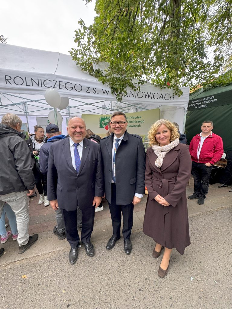 Grupa elegancko ubranych osób pozuje do zdjęcia przed zielonym namiotem wystawowym.