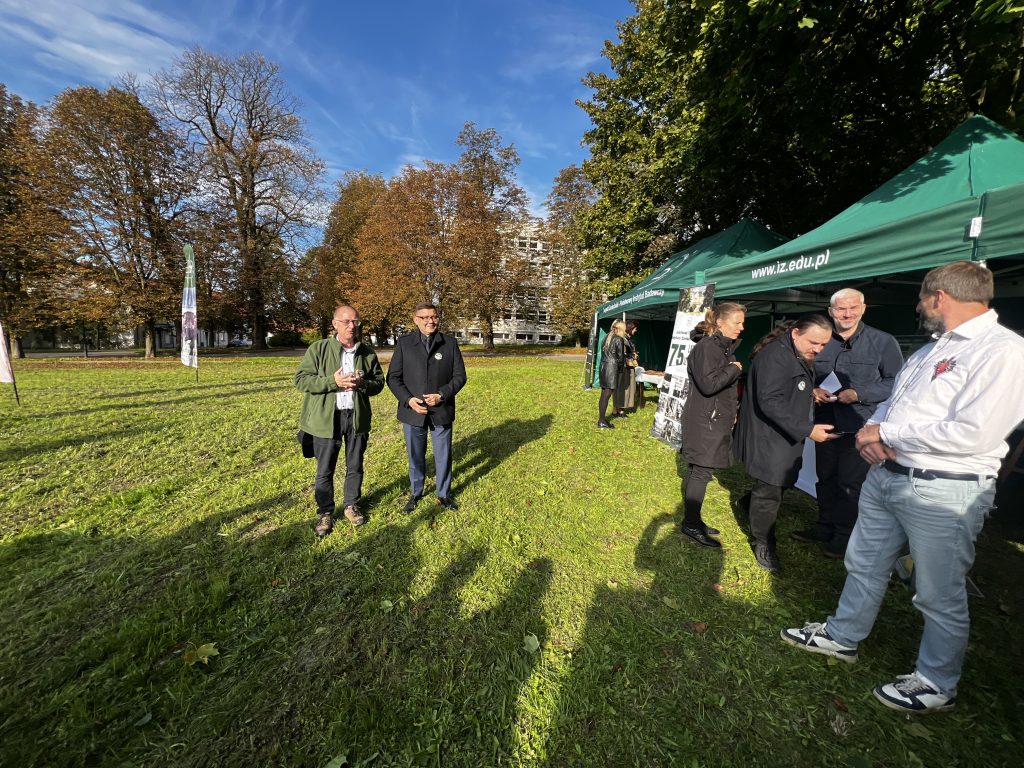 Grupa osób stoi przed namiotami Instytutu Zootechniki PIB podczas słonecznego dnia, w tle jesienne drzewa.