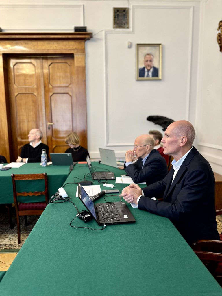 Mężczyzna w garniturze siedzący przy stole konferencyjnym z laptopem, w tle inni uczestnicy spotkania.