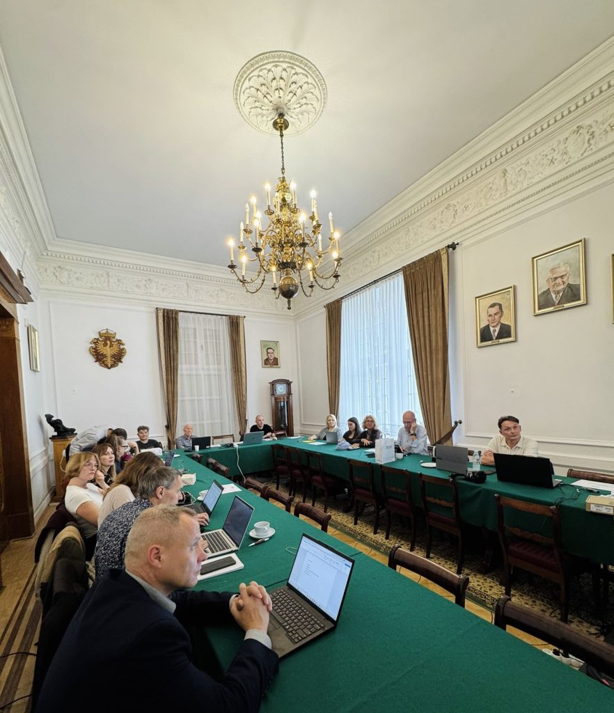 Uczestnicy spotkania siedzący przy długim stole z zielonym obrusem w eleganckiej sali z żyrandolem i portretami na ścianach.