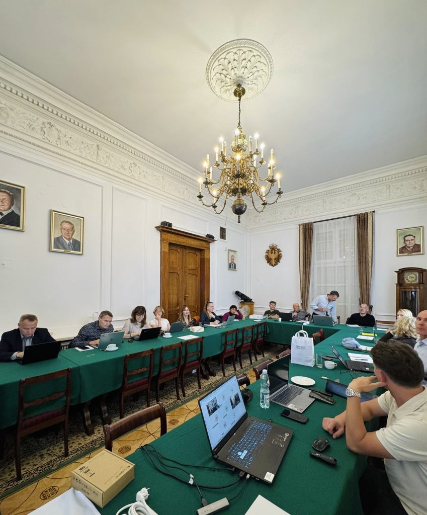 Widok na salę konferencyjną z uczestnikami pracującymi przy laptopach, w tle drewniane drzwi i zegar.