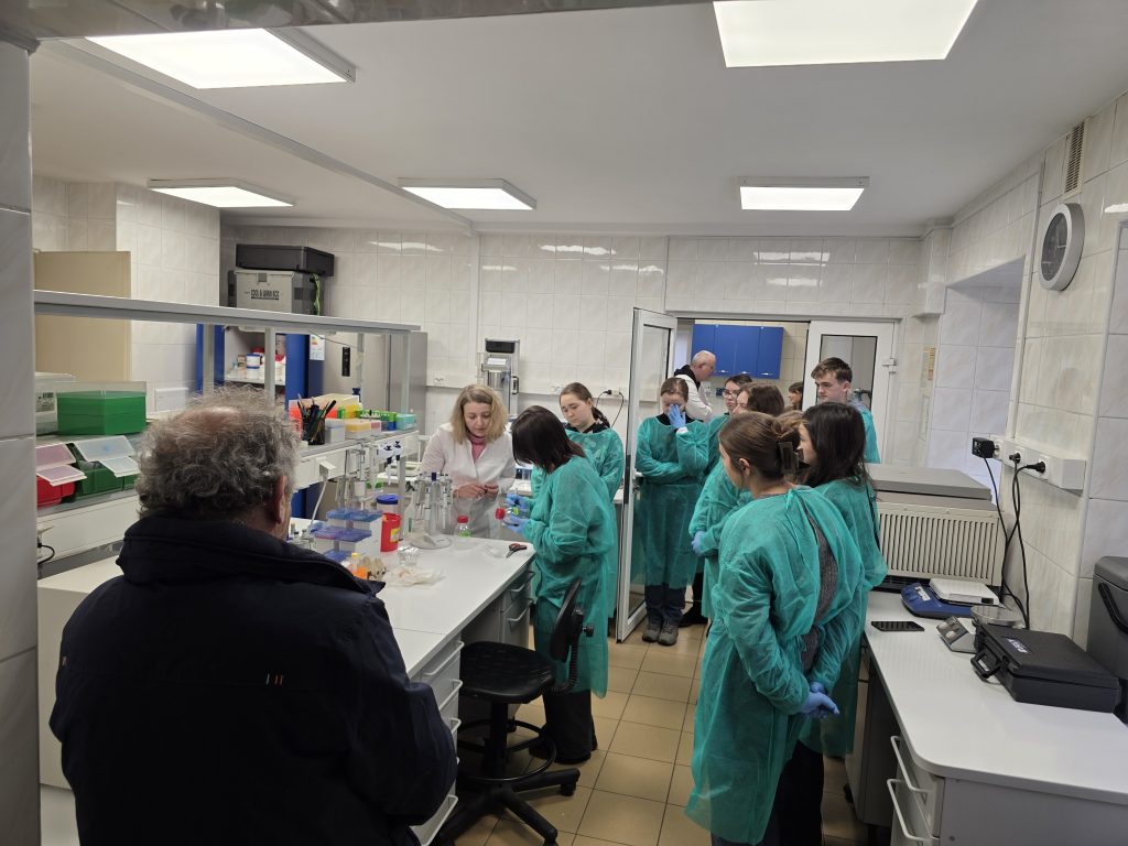 Grupa studentów w zielonych fartuchach ochronnych obserwuje prowadzącą podczas zajęć praktycznych w laboratorium.