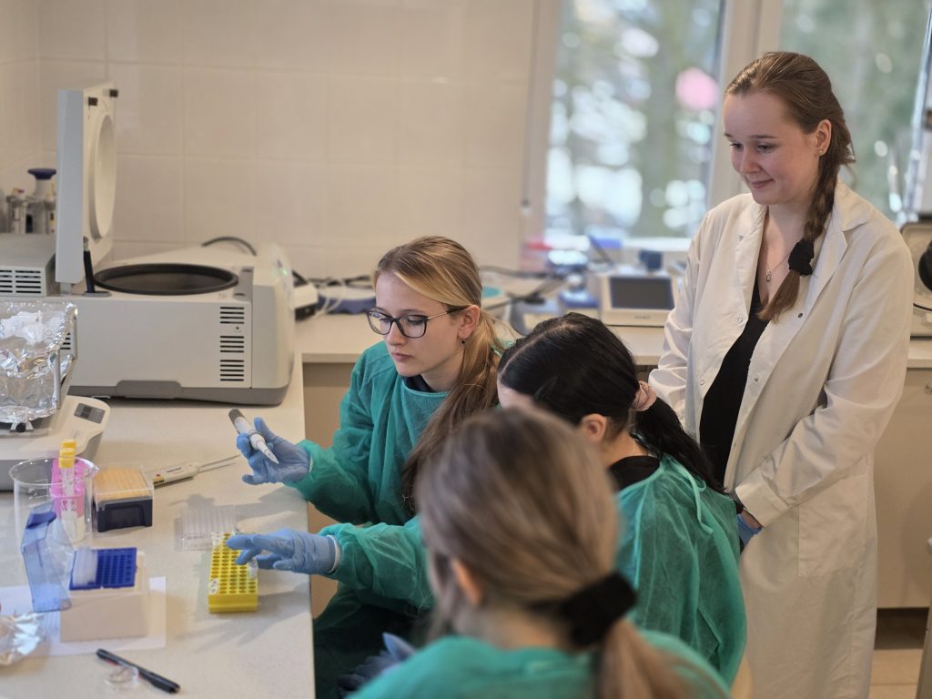 Studentki w fartuchach ochronnych wykonują ćwiczenia laboratoryjne, a prowadząca w białym kitlu nadzoruje pracę przy stanowisku.